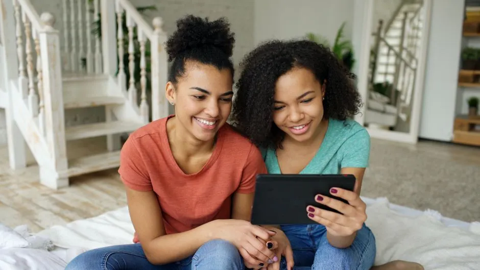 Two sisters sharing a tablet on a lounge room floor, both pointing at the same maths question